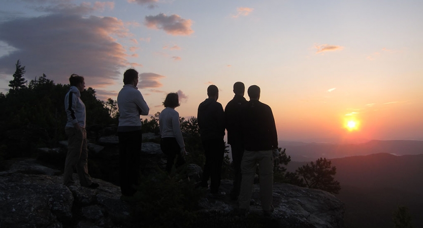 A group of people stand on an overlook and watch the sunset. They appear to be at high elevation in a mountainous landscape, and the sky is lit in soft hues of blue, yellow, orange, pink and purple.